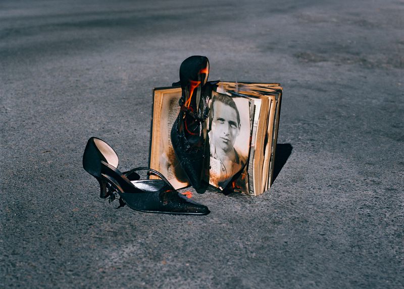 A book with a picture is ablaze, next to a black high-heel shoe on a grey pavement surface, in an outdoor daytime setting.
