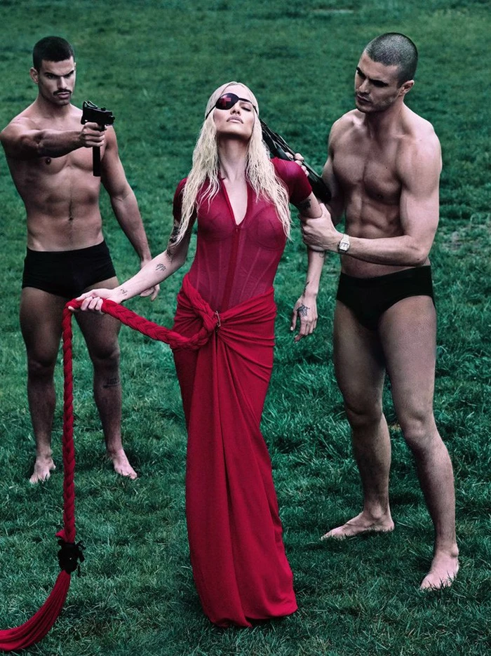 Gabbriette wearing a red gown and eye patch standing between two shirtless men on green grass for 032c by Steven Klein.