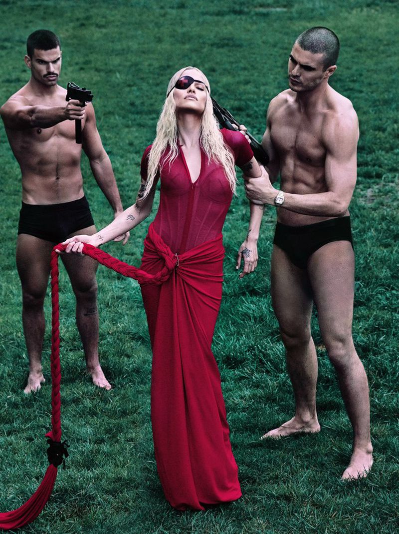 A woman in a red dress stands between two shirtless men holding guns, set on a green lawn, creating a visually striking and dramatic scene.