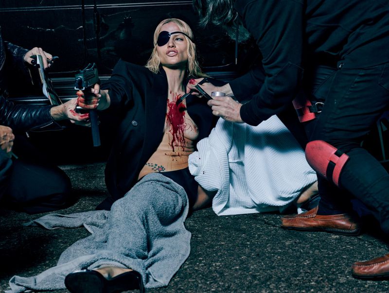 Dramatic scene with a woman lying on the ground, blood, a gun, and men standing around her in a dark setting.