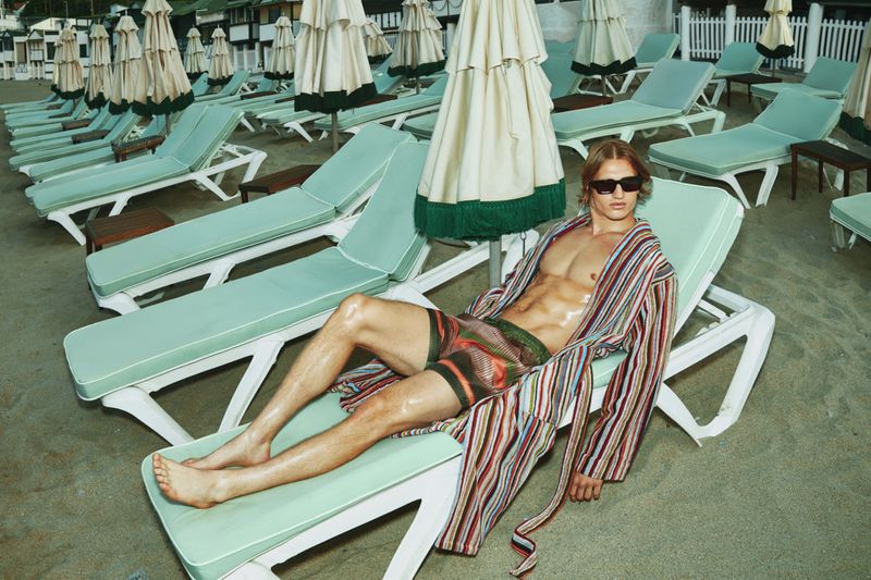 A blonde male model in sunglasses and striped robe is lying on a beach chair near a beach and white umbrellas in an outdoor fashion shot.