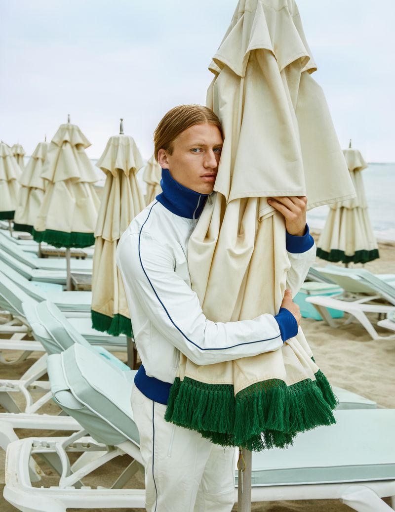 Young man poses on a beach, standing amid rows of closed beach umbrellas and lounge chairs. He wears a white and blue tracksuit.