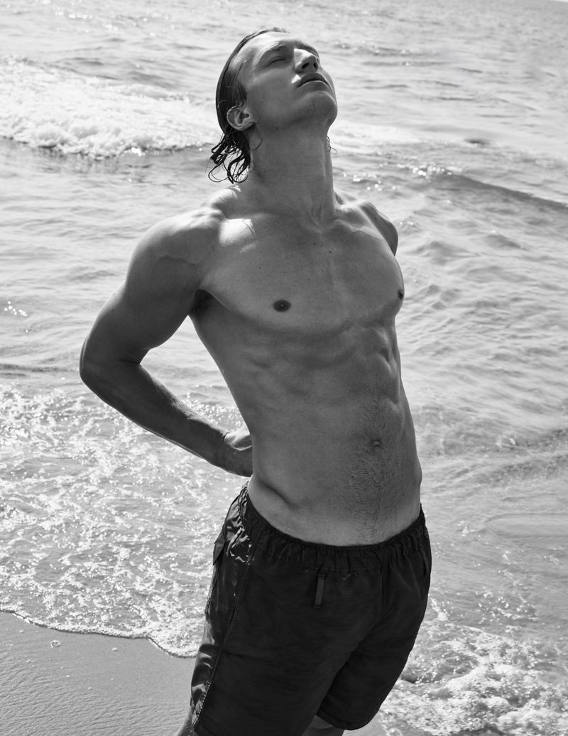 A shirtless adult male stands on a sandy beach with waves in the background in a black and white portrait.