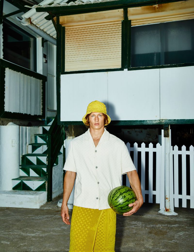 Young man in yellow bucket hat, white shirt, and wrap-around yellow skirt holds a watermelon in front of a white house and picket fence.