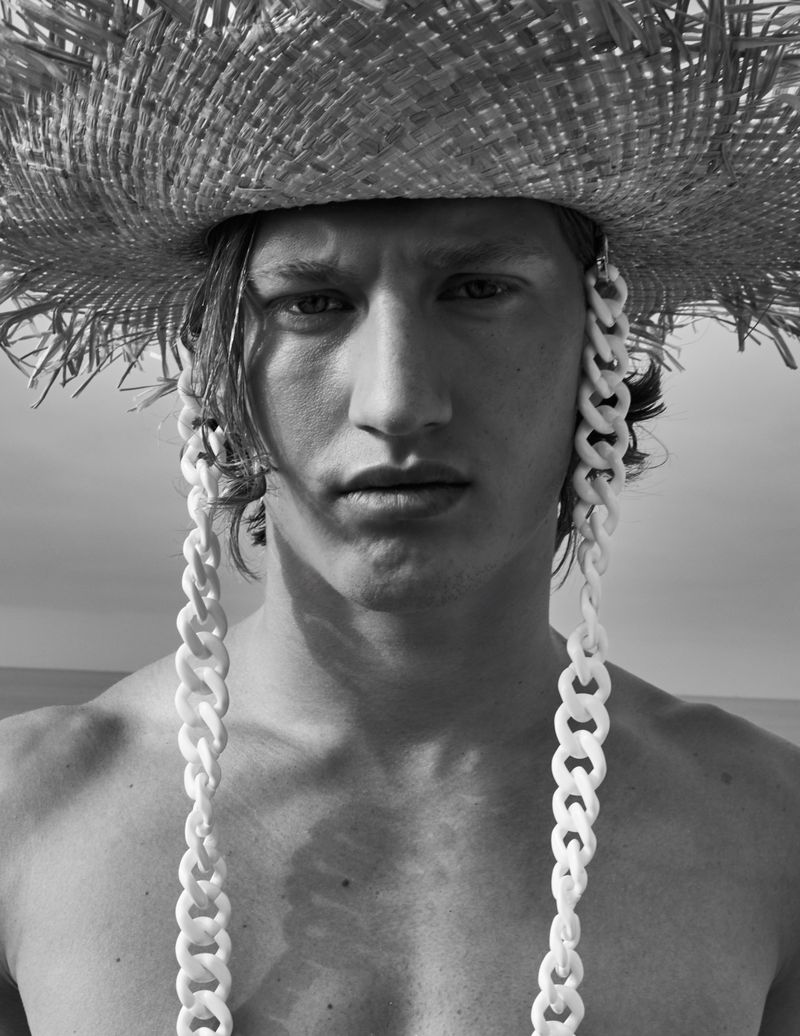 Black and white portrait of a shirtless man wearing a straw hat with long, white chain-like tassels, set against a bright sky.
