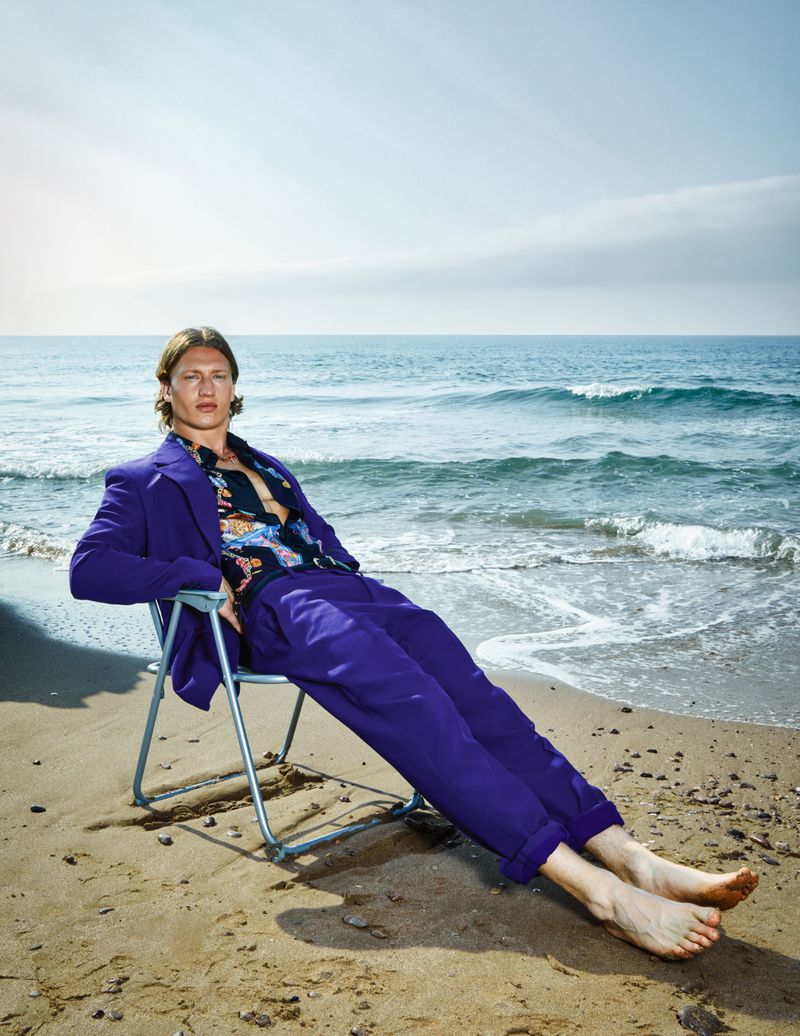 A fair-skinned adult man sits on a beach chair on a sandy beach with ocean and sky background. He wears a purple suit and patterned shirt.