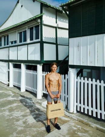 Shirtless male model stands outdoors with a Louis Vuitton tote bag, wearing striped shorts and black loafers, against a white building backdrop.