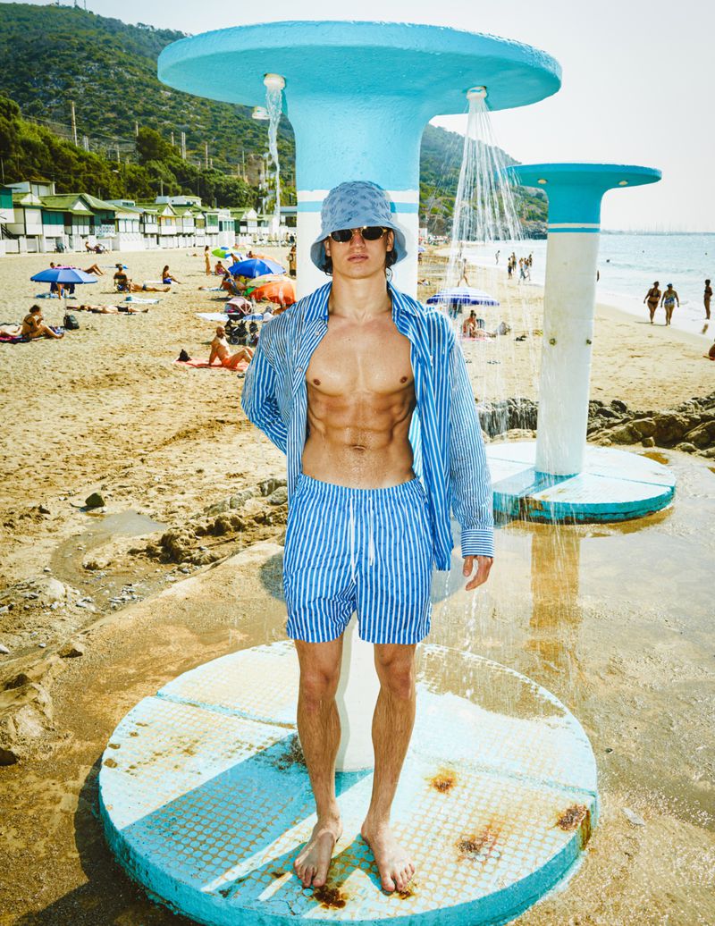 Man poses in blue striped shorts and shirt on a beach under a shower structure with a beach and trees in the background.