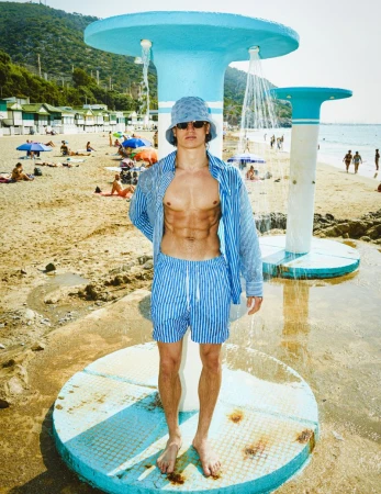 Man poses in blue striped shorts and shirt on a beach under a shower structure with a beach and trees in the background.