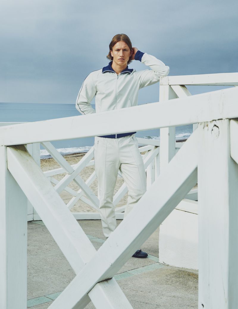 A fair-skinned man in a cream tracksuit with navy trim stands by a white railing on a beach against a cloudy sky, shot outdoors.