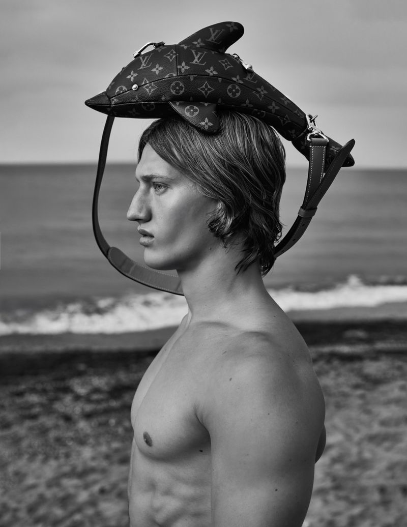 Black and white fashion portrait of shirtless male model wearing Louis Vuitton dolphin handbag on head near ocean.
