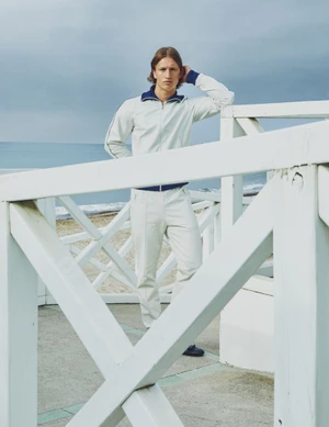 Male model wearing a white tracksuit standing behind a white wooden railing with an ocean beach background.