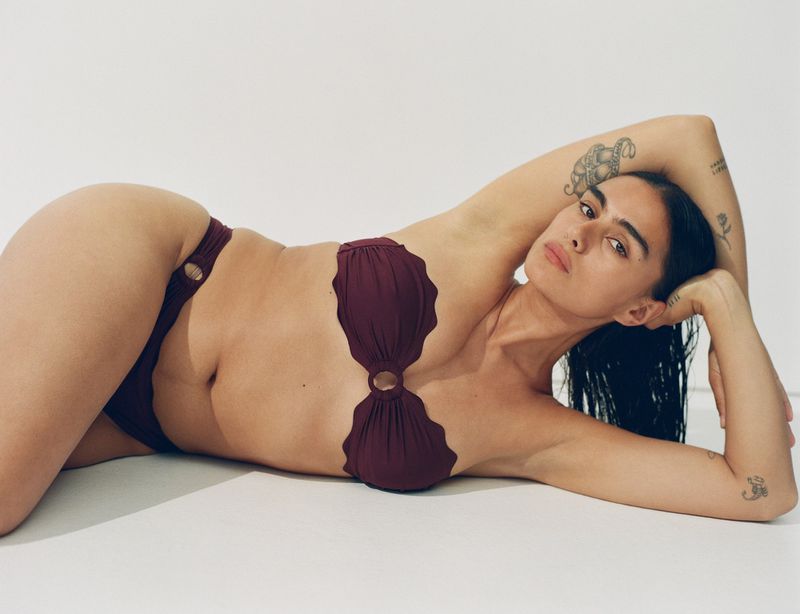 Woman in maroon two-piece swimsuit lying down against white backdrop, posing with hand behind head and looking at the camera.