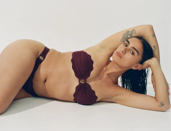 Woman in maroon two-piece swimsuit lying down against white backdrop, posing with hand behind head and looking at the camera.