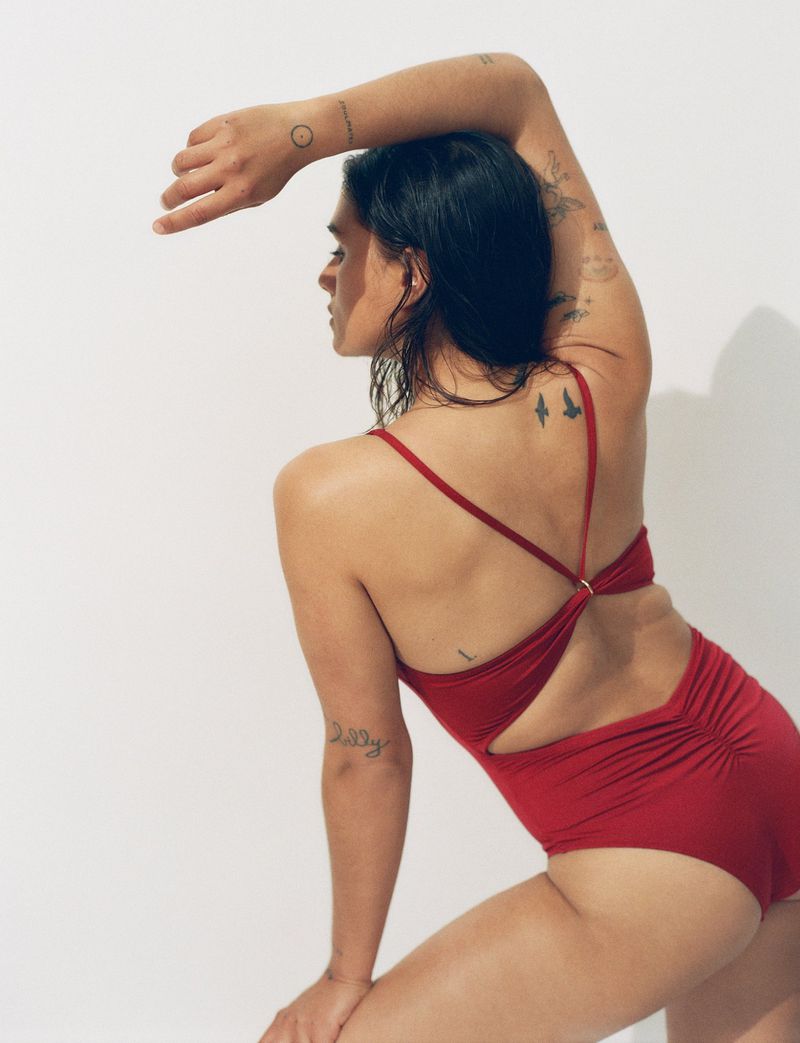 A brunette woman with tattoos poses indoors in a red one-piece swimsuit against a white background in a fashion style photograph.