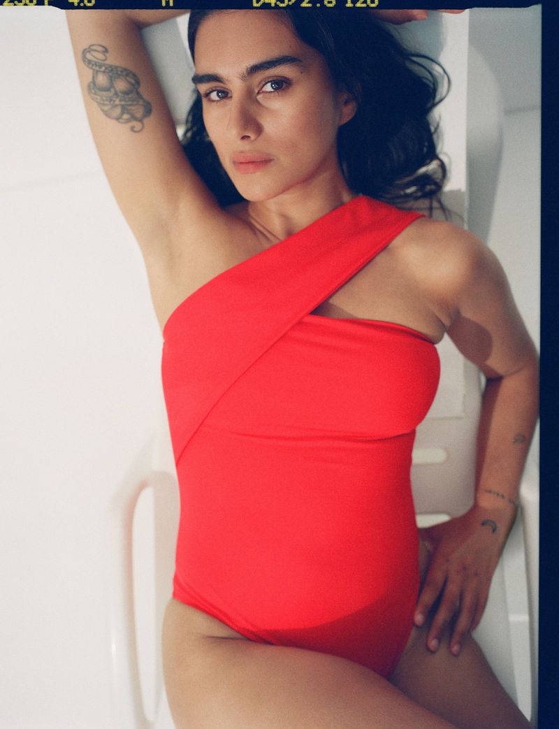Brunette woman in a red one-piece swimsuit poses in a white indoor setting, showcasing fashion, beauty, and lifestyle elements.