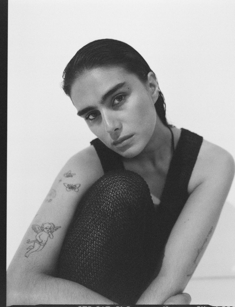 Monochrome portrait of a young adult female with short dark hair. She poses in a tank top with tattoos on her left arm and shoulder.