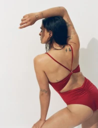 Model Jill Kortleve wearing a ruched red one-piece swimsuit with cross-back straps posing against a white studio background.