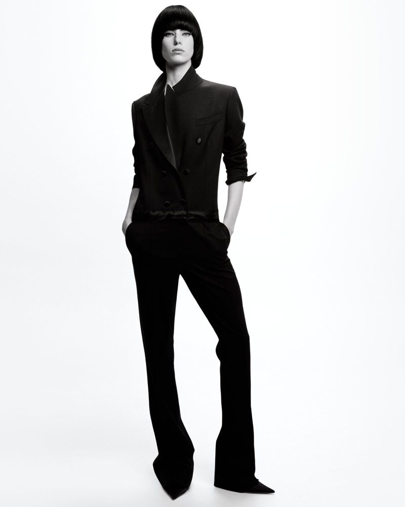 Monochrome studio shot of a woman with dark hair wearing a black double-breasted jacket and flared black trousers, minimalist style.