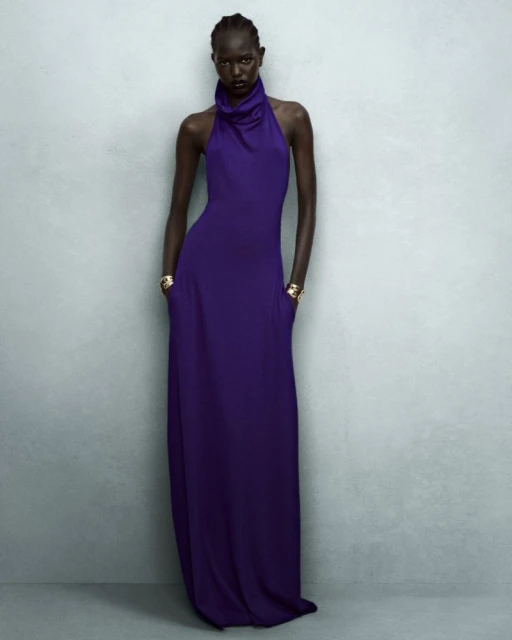 Elegant woman in a draped, floor-length halter dress strikes a pose against a neutral grey backdrop in a studio fashion shot.