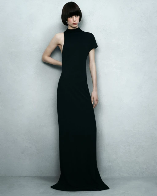 Elegant woman in a long black gown poses against a textured gray backdrop, fashion photography showcasing a sophisticated, minimalist aesthetic.