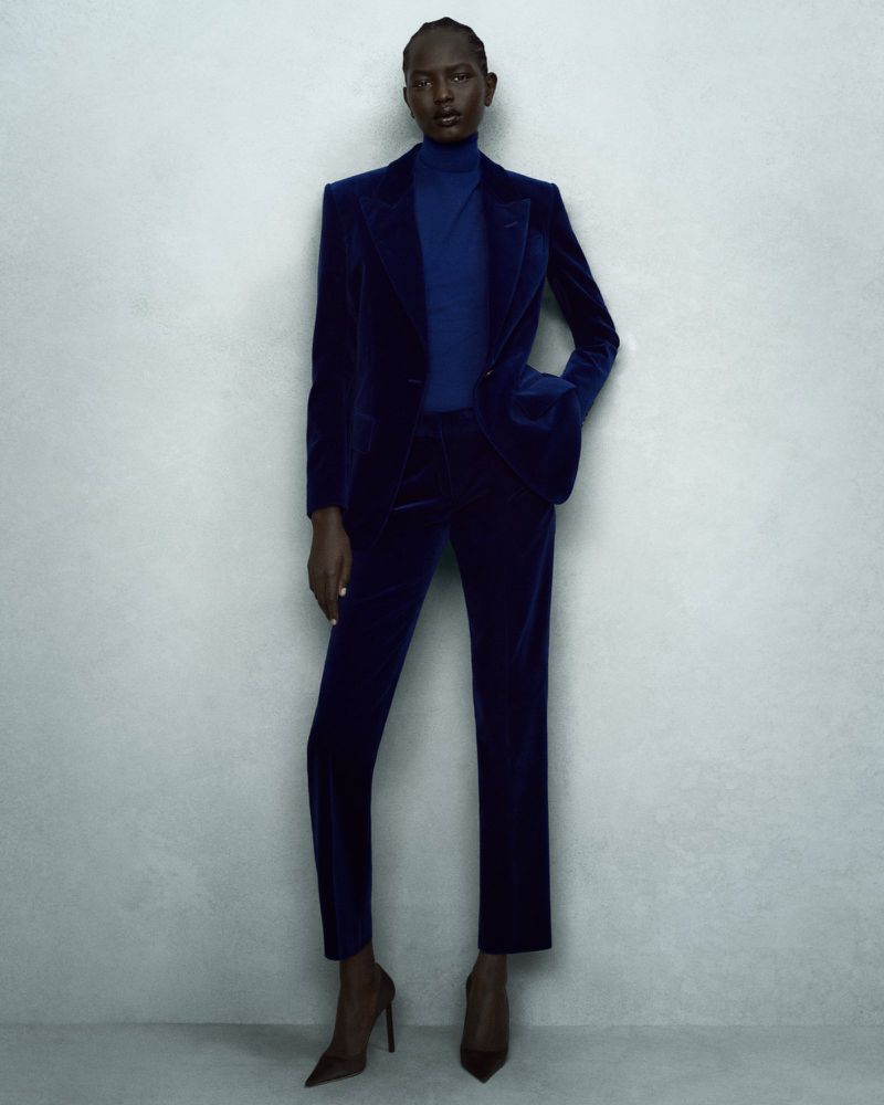 Woman wearing a blue velvet suit with a blazer and trousers, paired with high heels, standing against a neutral wall.