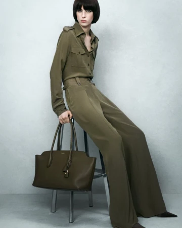 A woman models a tailored, army-green shirt and wide-leg pants, seated and holding a large leather tote bag against a plain backdrop.