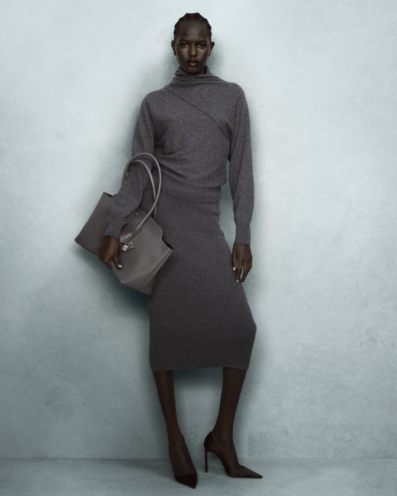 A slender woman in a grey turtleneck dress and heels holds a grey tote bag against a grey textured wall in a fashion studio.