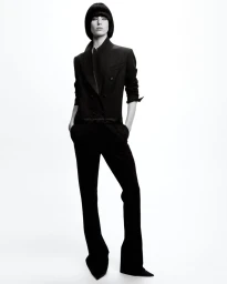 Model with a black bob haircut wearing a tailored black suit and pointed shoes against a bright white seamless background.