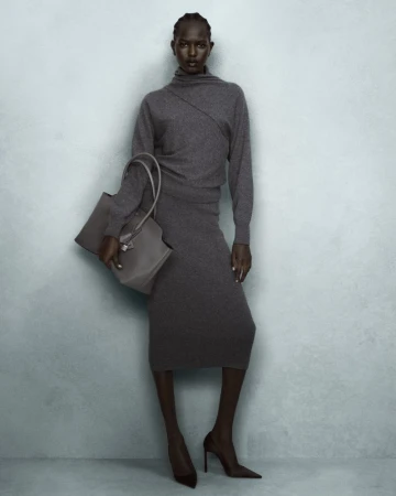 A Black woman in a grey knit sweater and matching midi skirt holding a grey tote bag against a textured grey wall.