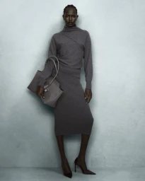 A Black woman in a grey knit sweater and matching midi skirt holding a grey tote bag against a textured grey wall.