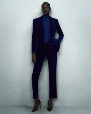 A Black female model wearing a tailored royal blue velvet suit and turtleneck with high heels against a textured grey studio backdrop.
