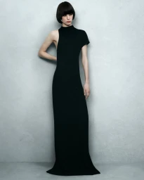 Model with dark bob hairstyle wearing a black sleeveless long dress against a grey textured studio backdrop.