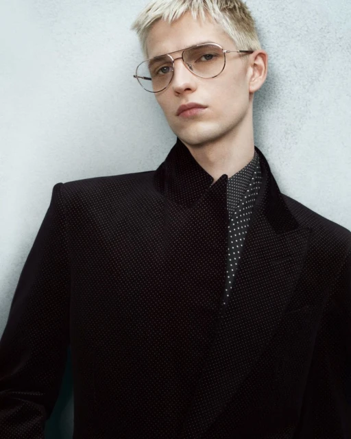 Close-up studio shot of a fair-skinned adult male model with platinum blonde hair wearing glasses and a polka dot blazer.