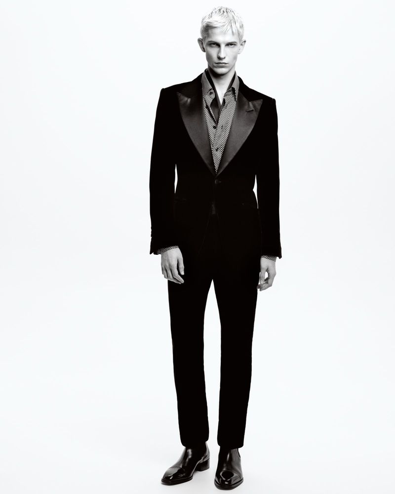 Monochrome portrait of a slender male fashion model in a dark tuxedo, posing in front of a plain white background in a studio setting.