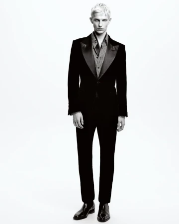 Monochrome portrait of a slender male fashion model in a dark tuxedo, posing in front of a plain white background in a studio setting.