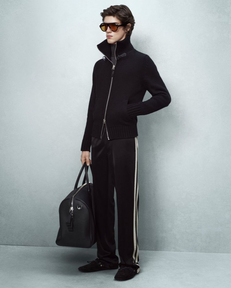 Adult male fashion model poses in dark lounge wear carrying a black duffel bag in a studio lit indoor setting on a grey background.