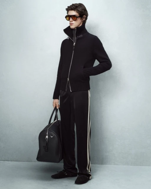 Adult male fashion model poses in dark lounge wear carrying a black duffel bag in a studio lit indoor setting on a grey background.