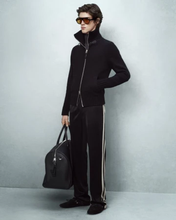 Adult male fashion model poses in dark lounge wear carrying a black duffel bag in a studio lit indoor setting on a grey background.
