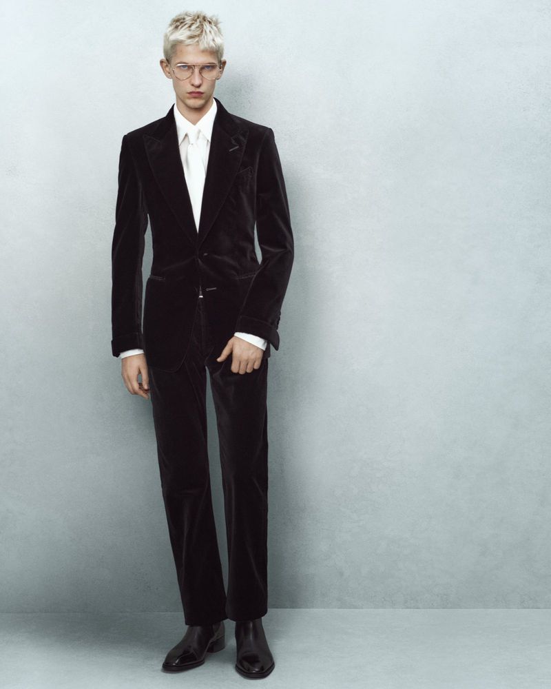 Male model in a black velvet suit and glasses posing against a grey wall in a studio, showcasing formal fashion and style.