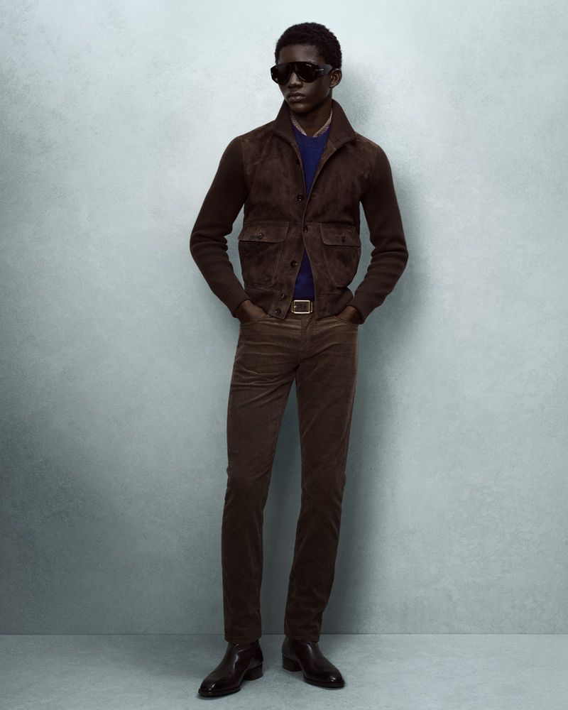 Full-length portrait of an African American man in a brown suede jacket, corduroy pants, sunglasses, and leather shoes against a neutral backdrop.