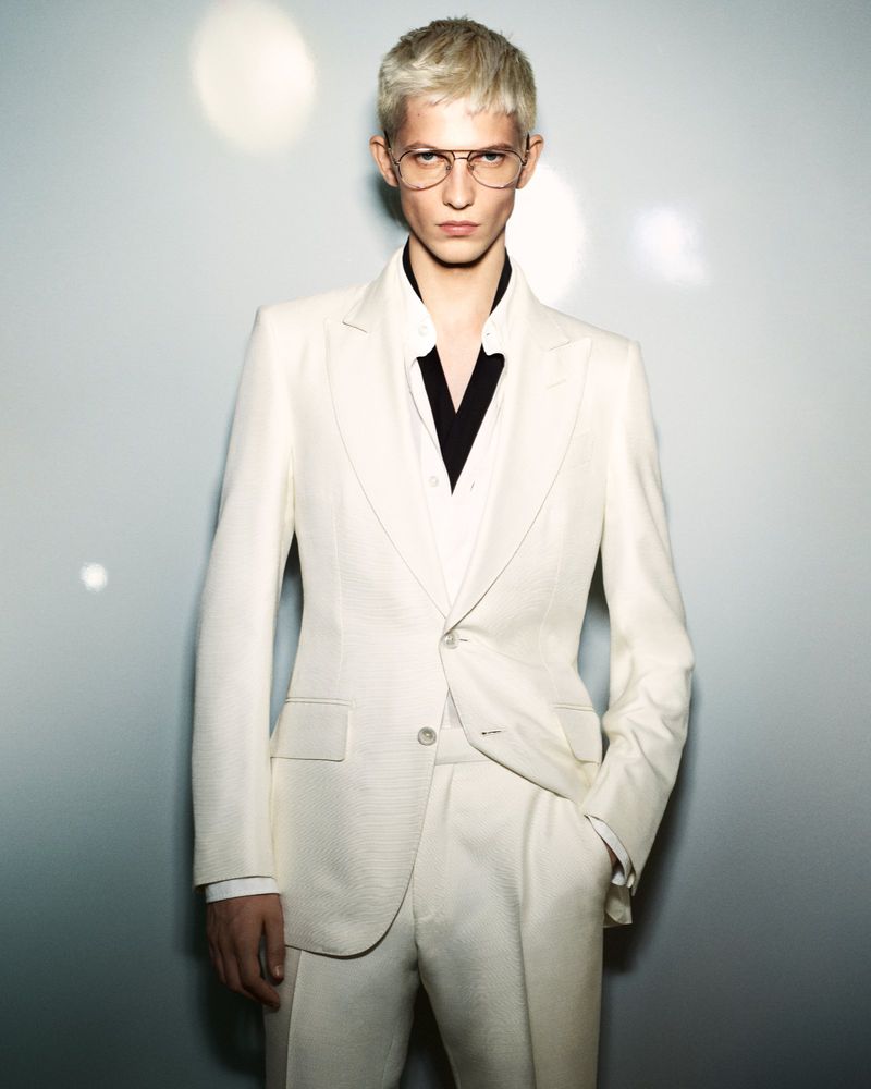 Blonde male model wearing a white suit and wire glasses against a grey studio backdrop