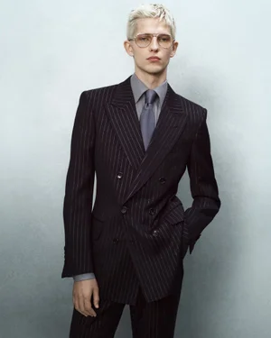 Blonde man wearing a dark pinstripe double-breasted suit and grey shirt against a pale wall.