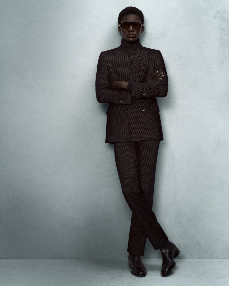 Black man wearing sunglasses and a dark suit standing with arms crossed against a textured grey studio wall