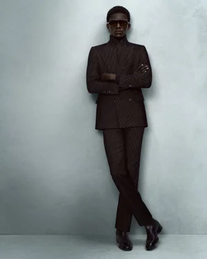 Black man wearing sunglasses and a dark suit standing with arms crossed against a textured grey studio wall