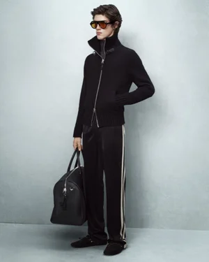 Male model wearing black zip-up jacket and sunglasses holding black leather duffel bag against grey studio background.