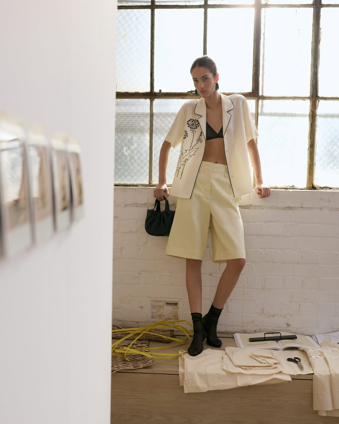 A fashionable woman poses near a sunlit window wearing a floral-embroidered top, matching shorts, black socks and boots, holding a small black purse.