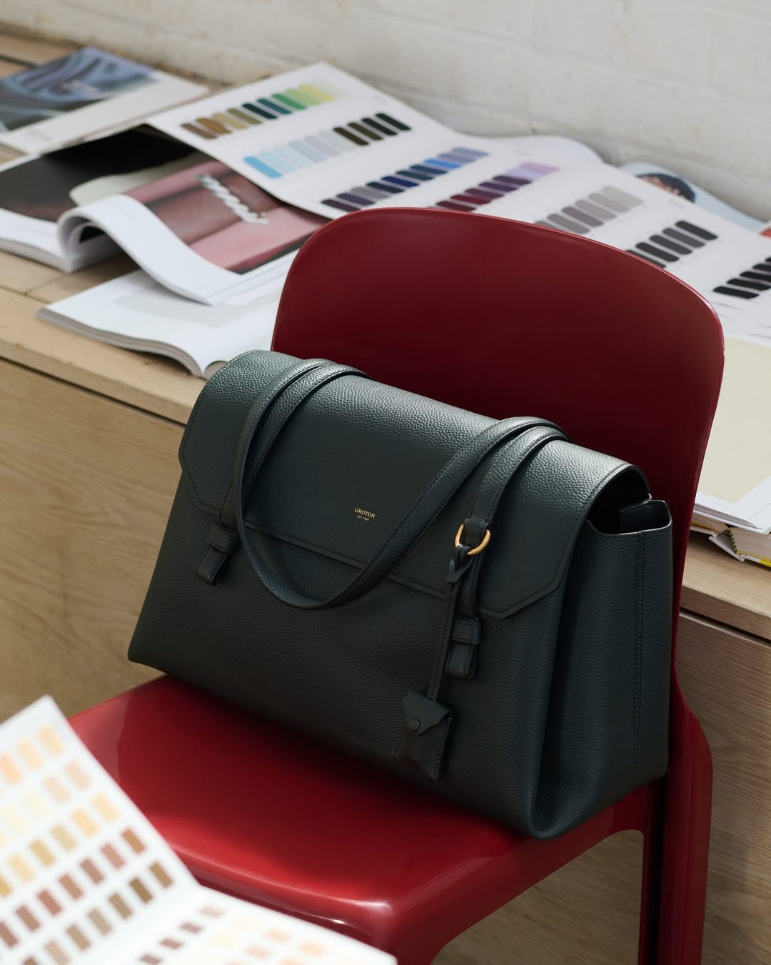 Elegant dark green leather handbag on a red chair, with color palettes and books in the background, suggesting a fashion or design context.