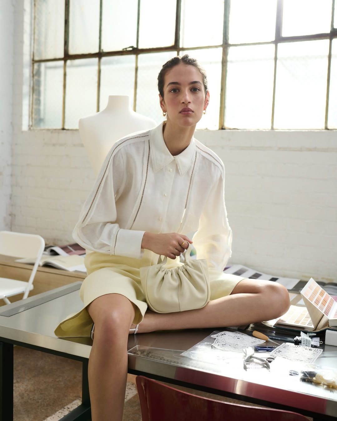 A young woman in a white linen blouse and yellow skirt sits on a metal table, holding a small off-white handbag in natural light.