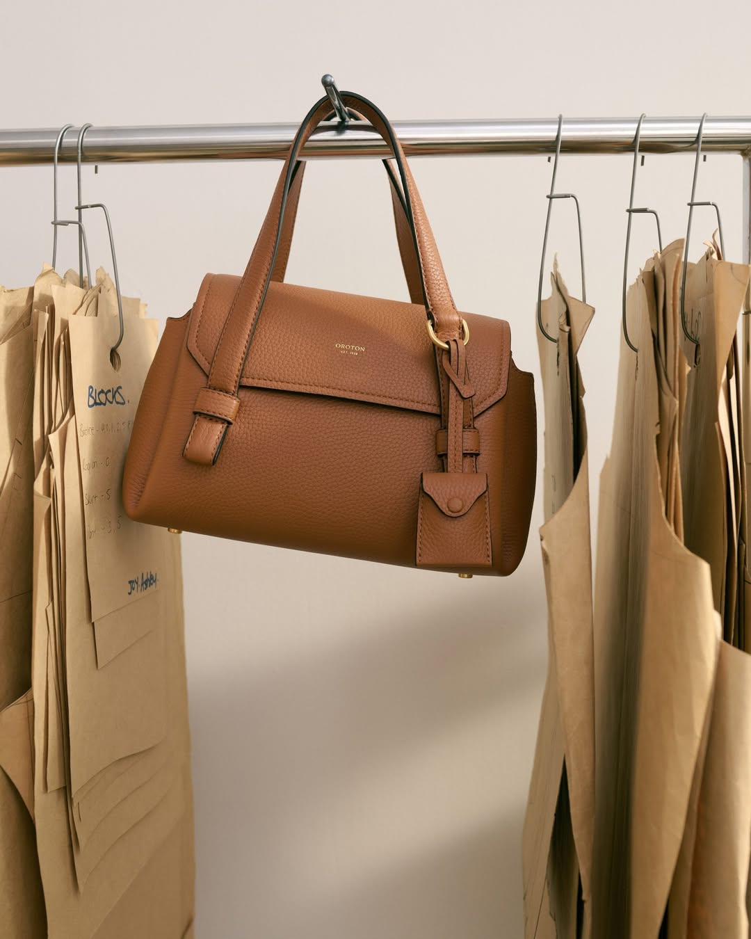 Brown leather handbag with gold hardware hangs on a metal rack near beige paper sewing patterns in a neutral indoor setting.